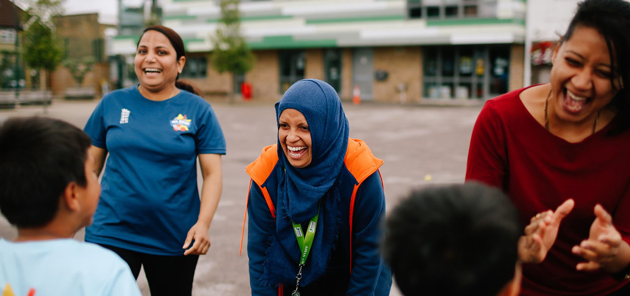 A group of female volunteers laugh as they interact with children on an outdoors playground